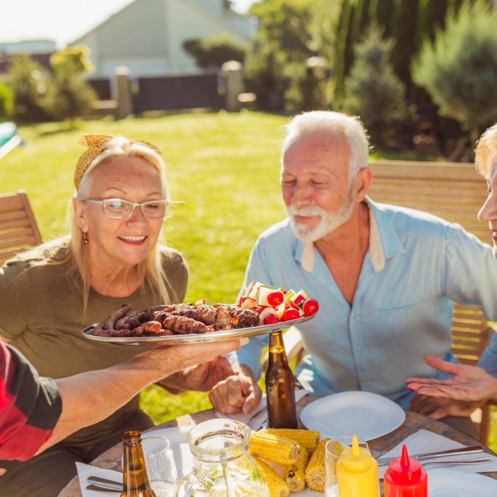 Group of cheerful senior friends having an outdoor lunch in the backyard, gathered around the table, host bringing food on a tray, offering it to guests